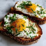 Two slices of savory ricotta toast topped with a sunny-side-up egg, fresh chives, and black pepper.