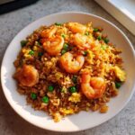 A close-up of a plate featuring a delicious shrimp fried rice recipe, garnished with large shrimp, green peas, and scrambled egg.