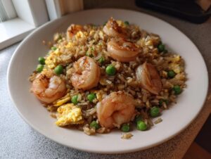 A close-up of a serving of shrimp fried rice recipe, featuring large shrimp, scrambled egg, and bright green peas.
