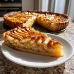 A close-up slice of a beautiful, glossy apple tart served on a white plate, with the rest of the apple tart visible behind it.