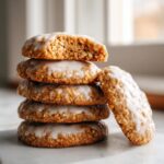 A stack of four soft iced oatmeal cookies, with the top one broken to show the texture, and one leaning against the side.