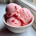 Three scoops of bright pink strawberry ice cream with visible fruit pieces served in a white bowl.