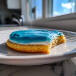 A close-up of a bitten sugar cookie topped with white and bright blue sugar cookie icing, sitting on a white plate.