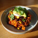 A close-up of a colorful sweet potato taco bowl featuring black beans, corn, topped with avocado, sour cream, and cilantro.