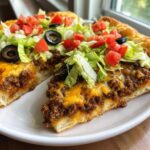 Close-up of a thick slice of taco pizza showing seasoned ground beef, melted cheese, lettuce, tomatoes, and black olives.