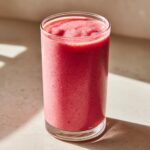 A tall glass filled with a thick, vibrant pink watermelon smoothie, sitting on a light surface with dramatic shadows.