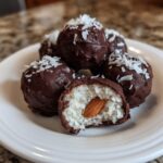 A stack of chocolate-covered almond joy truffles, one is cut open showing the coconut filling and whole almond inside.
