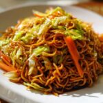 A close-up, appetizing shot of glossy, dark brown fried noodles mixed with bright green cabbage and orange carrot strips.