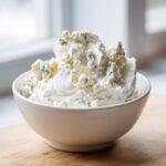 A close-up of a white bowl filled with chunky, homemade blue cheese dip, set on a wooden surface.