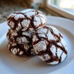 A stack of four rich chocolate crinkle cookies heavily dusted with powdered sugar, sitting on a white plate.