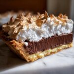 A close-up slice of chocolate meringue pie showing a golden crust, rich chocolate filling, and tall, toasted meringue topping.