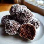 A pile of rich, dark chocolate bourbon balls rolled in shredded coconut, with one ball bitten open.