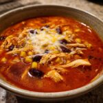 Close-up of a bowl of creamy chicken enchilada soup topped with shredded cheese, chicken, corn, and black beans.