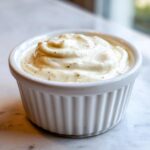 Close-up of creamy horseradish sauce, speckled with herbs, served in a small white fluted ramekin.