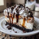 A rich slice of layered ice cream cake featuring a dark cookie crust, vanilla and chocolate layers, topped with whipped cream, sauces, and cookie crumbles.