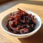 A small white bowl filled with deeply rich, dark brown caramelized onions, glistening under natural light.
