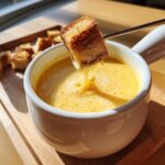 A piece of toasted bread is dipped into a white bowl of smooth, yellow cheese fondue recipe using a fondue fork.
