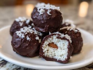 A stack of homemade almond joy truffles coated in dark chocolate and topped with shredded coconut, one is cut open showing the coconut filling and whole almond.