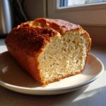 A close-up of a freshly baked, golden-brown beer bread loaf with a fluffy interior texture, sitting on a plate.