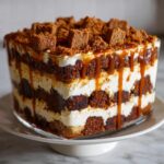 Close-up of a layered gingerbread trifle featuring dark cake cubes, white cream, and dripping caramel sauce.
