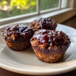 Three perfectly portioned mini meatloaf muffins topped with a shiny, dark red glaze, sitting on a light plate.
