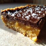 A close-up shot of a rich, layered graham cracker toffee bar topped with melted chocolate and flaky sea salt.