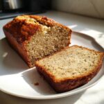 A golden-brown loaf of greek yogurt banana bread, partially sliced on a white plate.