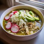 A vibrant bowl of green chicken pozole topped with shredded cheese, sliced radishes, avocado, and hominy.