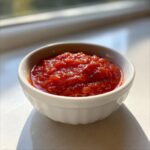 A small white bowl filled with vibrant, chunky homemade cocktail sauce, brightly lit by natural light.