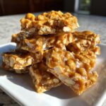 A stack of golden brown, glossy pieces of homemade peanut brittle recipe piled on a white plate.