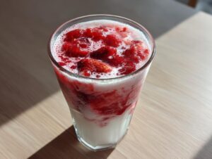 Close-up of a tall glass filled with layered homemade strawberry milk, featuring crushed strawberries and foam.