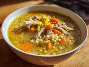 Close-up of a white bowl filled with Italian penicillin soup, featuring broth, small pasta pearls, shredded chicken, carrots, and lemon zest.