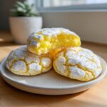 Three bright yellow lemon crinkle cookies coated in powdered sugar, with one cookie broken in half to show the soft interior.