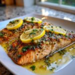Close-up of two pan-seared salmon recipe fillets topped with lemon slices and fresh parsley in butter sauce.