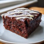 A close-up of a rich, dark square of gingerbread brownies topped with a white cream cheese icing drizzle.