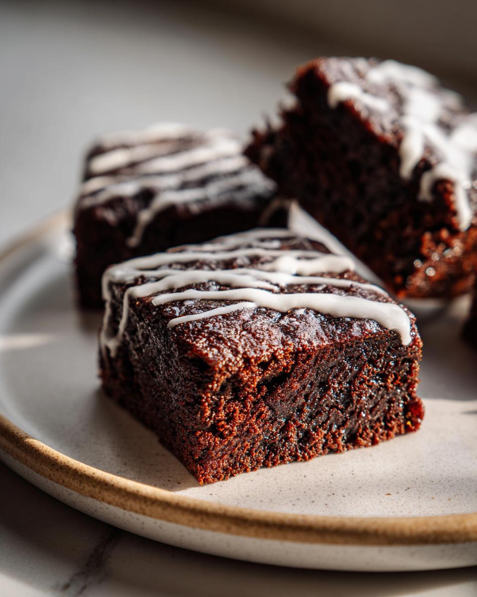Close-up of a fudgy, dark gingerbread brownie square topped with a white icing drizzle on a plate.