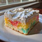 A moist slice of rainbow jello poke cake topped with thick white whipped cream, sitting on a white plate.