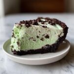 A thick slice of light green, creamy grasshopper pie with chocolate cookie chunks on a white plate.