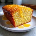 A close-up of a slice of moist orange juice cake topped with a thick, dripping orange glaze on a white plate.