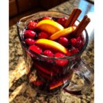 Close-up of a pitcher of deep red holiday sangria filled with fresh cranberries, orange slices, and cinnamon sticks.