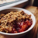 A close-up, appetizing photo of a single serving of homemade rhubarb crisp with a golden oat topping and bright red filling.