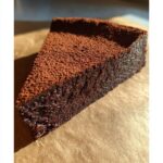 A close-up, rich slice of flourless chocolate torte, heavily dusted with cocoa powder, sitting on brown parchment paper.