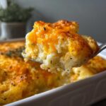 A spoonful of creamy, baked cheesy potato casserole topped with melted cheddar cheese being lifted from the dish.