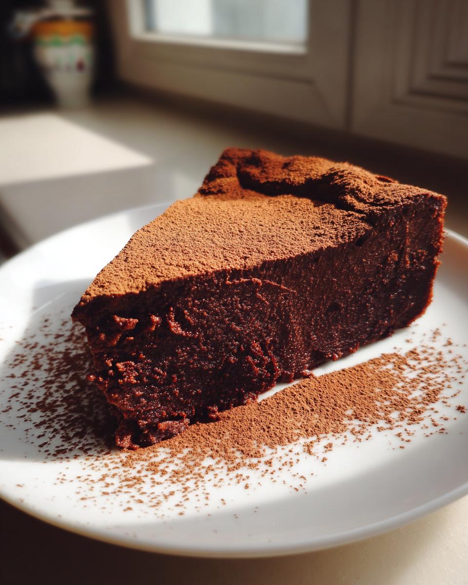 A decadent slice of flourless chocolate torte dusted heavily with cocoa powder, served on a white plate.