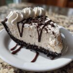 A decadent slice of no-bake ice cream pie with a dark cookie crust, topped with whipped cream and chocolate syrup.