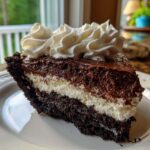 A decadent slice of layered mud pie featuring a dark crust, white cream layer, chocolate mousse, and topped with whipped cream.