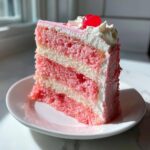A tall slice of pink and white layered cherry cake topped with whipped cream and a maraschino cherry.