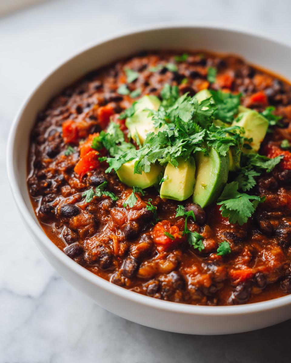 Close-up of a bowl of rich, smoky black bean chili topped with fresh avocado slices and cilantro.