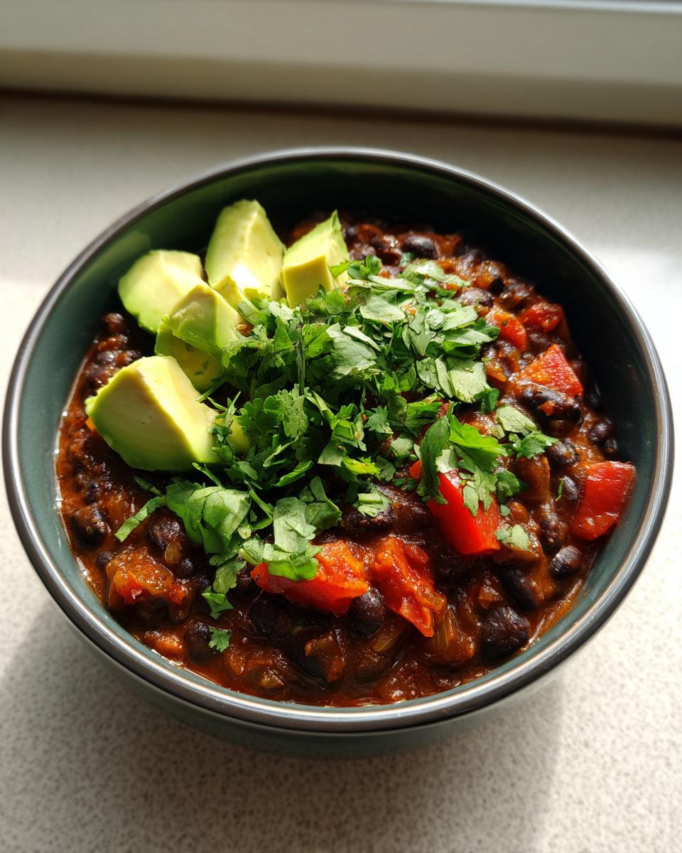 A close-up of a bowl of smoky black bean chili topped with fresh avocado slices and chopped cilantro.