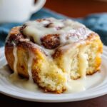 Close-up of a soft, gooey cinnamon roll with heavy cream icing dripping down the sides onto a white plate.